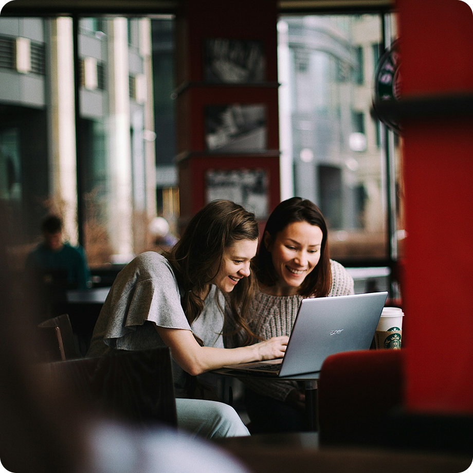 Dos mujeres sonríen mientras usan un portátil en una cafetería, compartiendo un momento de trabajo o estudio.